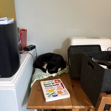 A black and white tuxedo cat on a table, next to a sewing book, with a hi fi on one side and a sewing machine on the other